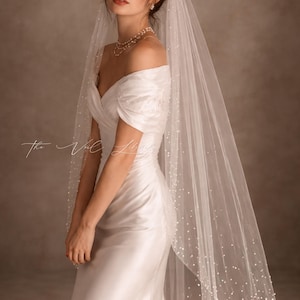Könnte beinhalten: Weißes Satin-Brautkleid mit schulterfreiem Ausschnitt. Ein durchsichtiger Schleier ist mit kleinen, verstreuten Perlen verziert. Der Text "The Veil Library" ist kursiv auf der linken Seite des Bildes geschrieben.