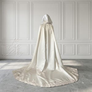May include: A cream-colored hooded cape with a long train, likely for a wedding or formal event. The cape is made of a satin-like material and features a delicate lace trim along the hem of the train. The hood is up, and the cape drapes elegantly.