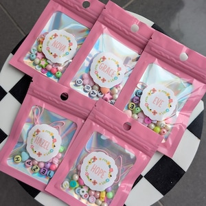 May include: Five pink holographic pouches containing assorted colourful beads and letter beads spelling the names Nova, Grace, Eve, Hazel, and Hope. Each pouch features a circular white label with a floral border and the name printed in the centre.