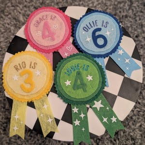 May include: Four circular felt birthday badges in pink, blue, yellow, and green. Each badge features a scalloped edge, a central number, and text including 'Grace is 4', 'Ollie is 6', 'Rio is 3', and 'Louie is 4'. Each badge has two hanging ribbons.