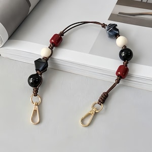 May include: A decorative handbag strap featuring a series of colorful beads in black, white, red, and navy blue. The beads are strung on a brown leather cord and attached to gold-colored clasps.
