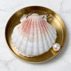 以下が含まれることがあります： 金色の丸いお皿に、白とピンクのホタテ貝と真珠が置かれています。ホタテ貝は扇形で、ピンクと白の縞模様があります。お皿は滑らかで光沢のある表面をしています。