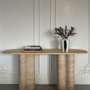 May include: A light beige travertine console table with a curved top and two cylindrical, fluted legs. A stack of books and a silver vase with dark branches sit on the table. The table is against a white wall.