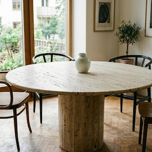 May include: A round dining table with a light-colored stone top and a cylindrical base. Four chairs surround the table, including a rattan armchair and three wooden chairs. A white vase sits on the table. The room has large windows and hardwood floors.