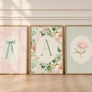 Puede incluir: Tres láminas decorativas enmarcadas. La lámina de la izquierda muestra un lazo azul claro sobre un fondo de rayas rosas. La central muestra la letra A sobre un estampado floral. La de la derecha presenta una flor rosa sobre un fondo de rayas verde claro.