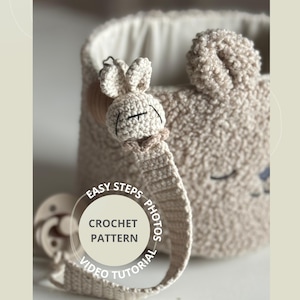 May include: A beige crochet baby basket with a matching pacifier clip and a plush bunny face. The basket has a soft, textured appearance. The pacifier clip has a bunny head. The text on the image says "EASY STEPS PHOTOS CROCHET PATTERN VIDEO TUTORIAL".