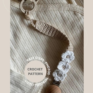 Op de afbeelding: Een gehaakte speenclip in beige met witte bloemaccenten. De clip heeft een houten sluiting en een ronde speenhouder. De afbeelding bevat de tekst "Easy Steps Photos Crochet Pattern Video Tutorial".