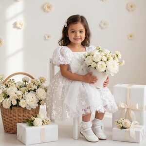 May include: A young girl in a white sequined dress with a bow, holding a white pot of white roses. A wicker basket and wrapped gifts with white roses are also in the image. The background is white with floral decorations.