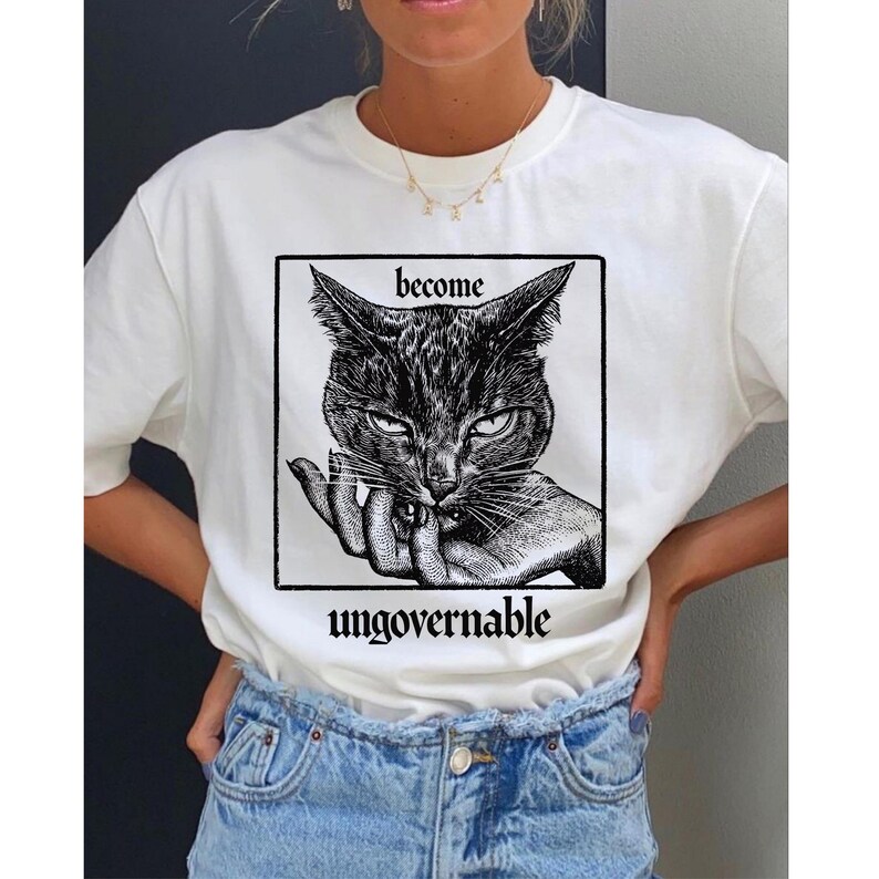 Puede incluir: Camiseta blanca con un gr&aacute;fico en blanco y negro de la cara de un gato dentro de un marco cuadrado. Las palabras "become" y "ungovernable" est&aacute;n impresas encima y debajo de la ilustraci&oacute;n del gato. La camiseta es de material suave.