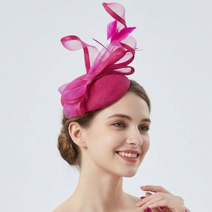 Puede incluir: Un fascinador fucsia vibrante, con un lazo y detalles de tela transparente. El tocado presenta una base pequeña y redondeada, adornada con plumas y cintas. Este elegante accesorio es adecuado para ocasiones especiales.