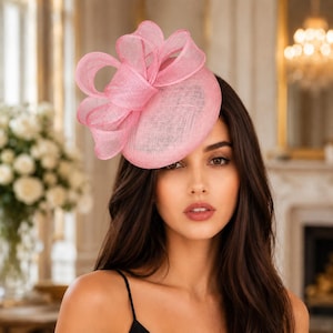Op de afbeelding: Een lichtroze fascinator met een ronde, getextureerde basis versierd met grote, gestructureerde lussen van bijpassende transparante stof. Het accessoire is gemaakt van een geweven materiaal met een zichtbaar gaasachtig patroon.