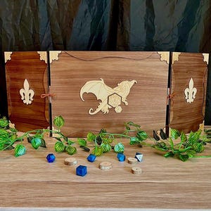 Könnte beinhalten: Ein hölzerner Tischspielbildschirm mit einem lasergeschnittenen Drachendesign und Würfeln. Der Bildschirm ist braun mit goldenen Akzenten und Fleur-de-Lis-Details. Blaue Würfel und Holzscheiben sind davor verstreut.
