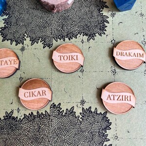 Könnte beinhalten: Runde Holzspielsteine mit den eingravierten Namen "ELTAYE", "TOIKI", "DRAKAIM", "CIKAR" und "ATZIRI". Jeder Spielstein hat ein bannerartiges Design. Sie sind auf einer kartenartigen Oberfläche angeordnet.