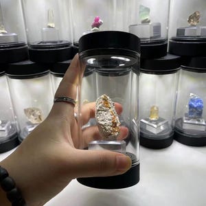 May include: A rough, textured mineral specimen with white and orange-brown coloration, displayed inside a clear cylindrical plastic container with a black lid and base, positioned on a small transparent stand within the enclosure.
