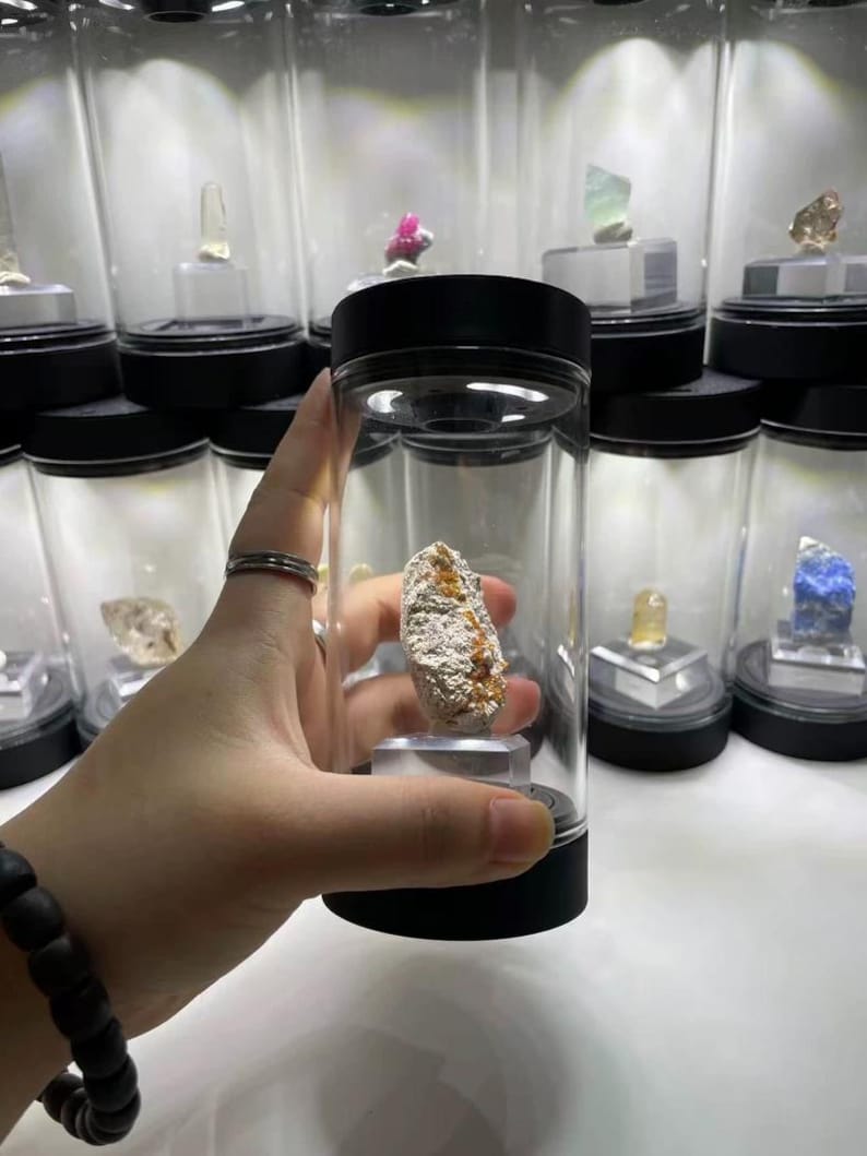 May include: A rough, textured mineral specimen with white and orange-brown coloration, displayed inside a clear cylindrical plastic container with a black lid and base, positioned on a small transparent stand within the enclosure.