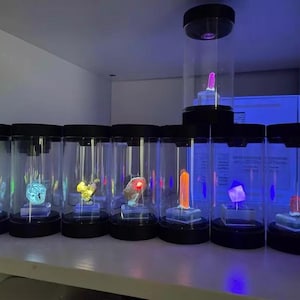 May include: A collection of eight clear cylindrical display cases with black lids and bases, each containing a single mineral specimen illuminated by ultraviolet light, showcasing vibrant fluorescent colors including neon green, blue, yellow, and orange.