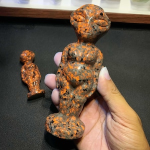 May include: Two polished stone figurines carved into the shape of an alien figure with a large head, almond-shaped eyes, and a rounded body. The stone features a speckled pattern of vibrant orange and dark grey colours throughout the surface.