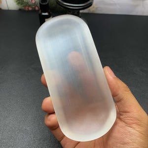 May include: A smooth, elongated, oval-shaped selenite crystal slab with a translucent, milky white appearance and a fibrous, satin-like texture. The crystal has rounded edges and a flat surface, displaying natural light-reflecting properties.