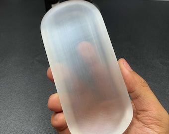 May include: A smooth, elongated, oval-shaped selenite crystal slab with a translucent, milky white appearance and a fibrous, satin-like texture. The crystal has rounded edges and a flat surface, displaying natural light-reflecting properties.