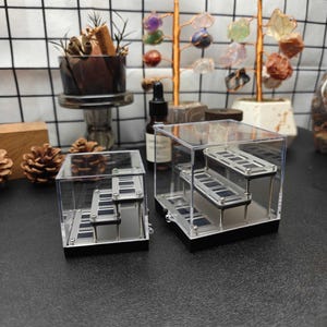 May include: Two clear acrylic display cases with black bases, each containing a tiered staircase structure. The internal stairs feature dark rectangular panels arranged in a step-like formation, supported by thin metal posts within the transparent boxes.