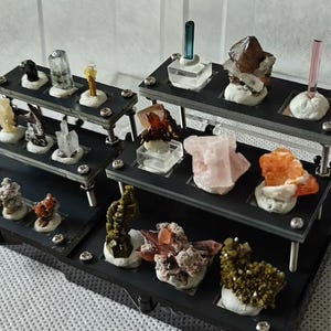 May include: A tiered black display stand holding a collection of various raw mineral specimens. The crystals feature diverse colors including pink, orange, green, brown, and clear, each mounted on a small white base within the stand's square cutouts.