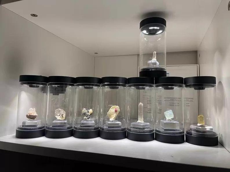 May include: A collection of eight cylindrical display cases with black lids and bases, each containing a unique mineral specimen on a small square stand. Seven cases are arranged in a row, with one additional case elevated on top.