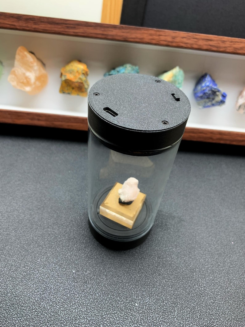 May include: A cylindrical display case with a black top and base, featuring a clear glass body. Inside, a small, pale pink mineral specimen sits atop a square, tan-colored pedestal. The black top includes a small port and visible screw heads.
