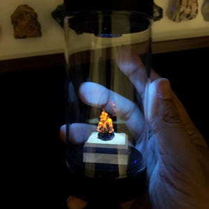 May include: A clear cylindrical display container with a black lid and base, featuring a small, jagged mineral specimen that glows with a bright orange fluorescent light. The specimen sits on a square white pedestal inside the transparent tube.