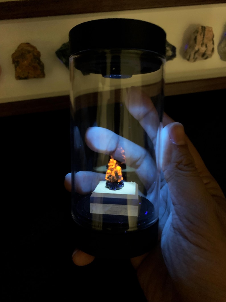 May include: A clear cylindrical display container with a black lid and base, featuring a small, jagged mineral specimen that glows with a bright orange fluorescent light. The specimen sits on a square white pedestal inside the transparent tube.