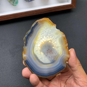 May include: A polished agate slice featuring concentric bands of translucent white, grey, and amber colors. The center displays a crystalline texture with a bright, glowing appearance, surrounded by a dark blue-grey ring and an outer amber edge.