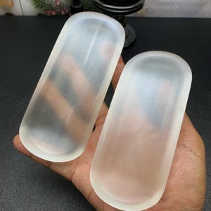 May include: Two elongated, oval-shaped trays carved from translucent, milky white selenite crystal. Each tray features a smooth, polished surface with rounded edges and a shallow, concave centre, showcasing the natural fibrous texture of the stone.