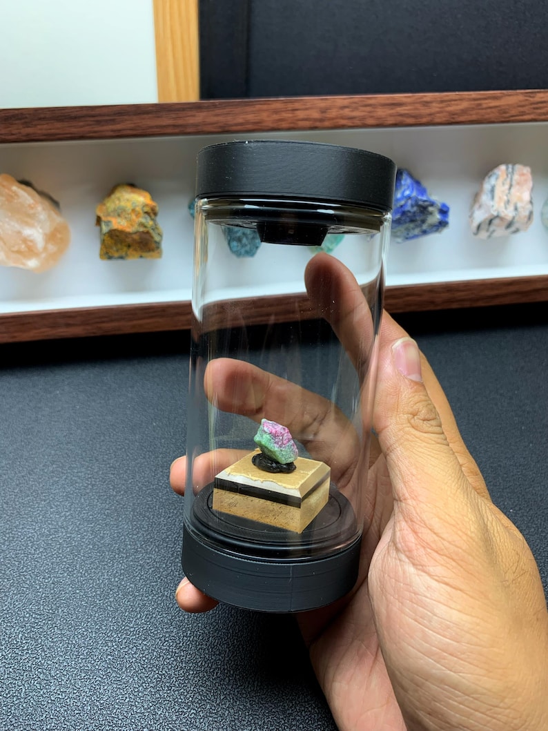 May include: A small, raw, pink and green mineral specimen sits on a square, layered tan and black base inside a clear, cylindrical glass display container with a black lid and base. The container is held upright, showcasing the crystal inside.