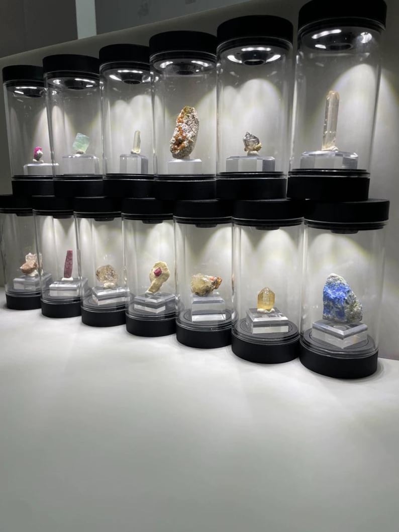 May include: A collection of twelve clear cylindrical display cases with black lids and bases, each containing a unique mineral specimen mounted on a small square stand. The cases are arranged in two rows, illuminated by internal top-mounted lights.