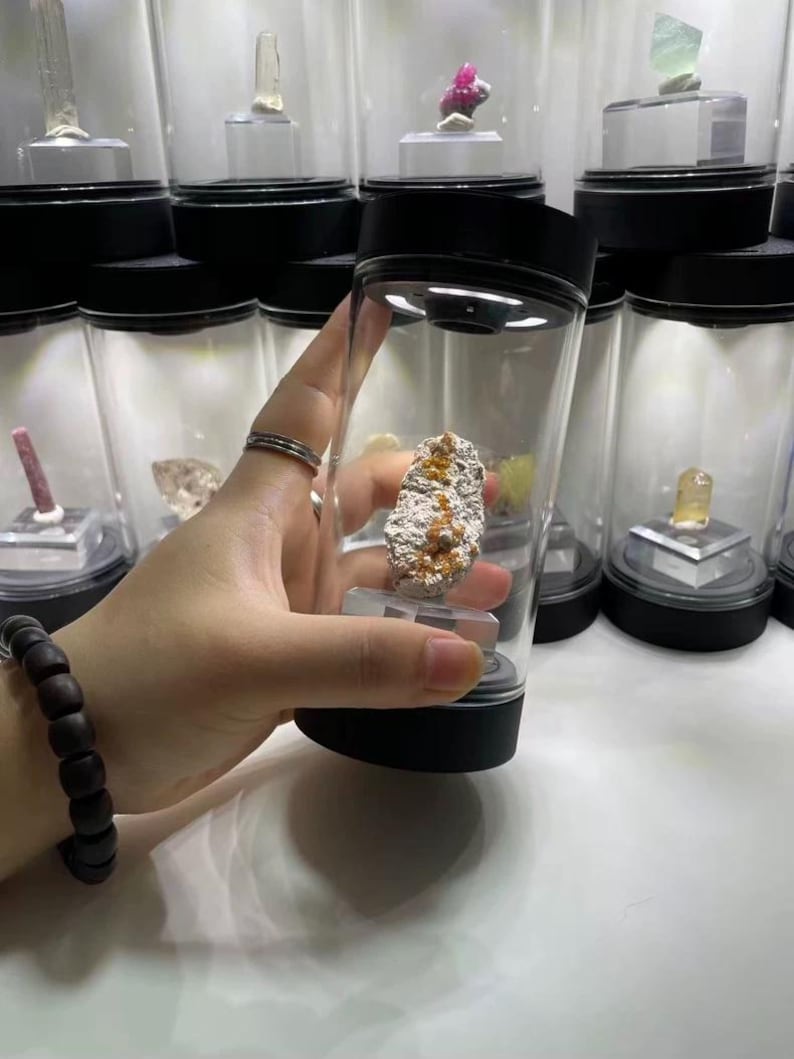 May include: A clear cylindrical display case with a black base and lid, featuring an integrated LED light at the top. Inside the container, a rough, textured mineral specimen with orange and white crystalline deposits sits on a small clear stand.