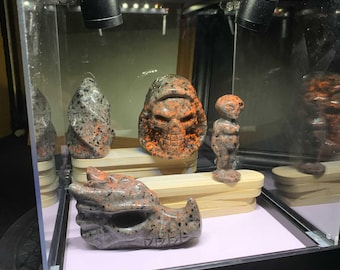 May include: A collection of four carved stone figurines made of speckled orange and grey granite. The set includes a dragon head, a human skull, a small humanoid figure, and a flame-shaped piece, all displayed inside a glass case with lights.