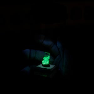 May include: A small, rough-textured mineral specimen emitting a bright, neon green glow in a dark environment. The mineral is mounted on a square, light-coloured base, creating a stark contrast against the surrounding black background.