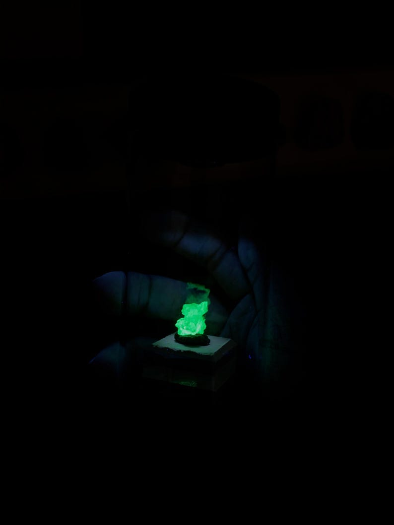 May include: A small, rough-textured mineral specimen emitting a bright, neon green glow in a dark environment. The mineral is mounted on a square, light-coloured base, creating a stark contrast against the surrounding black background.