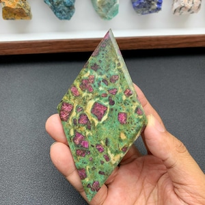 May include: A diamond-shaped polished stone featuring a vibrant green matrix speckled with deep magenta and red crystalline inclusions. The surface displays a smooth, flat texture with distinct color variations throughout the mineral specimen.