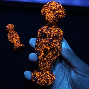 May include: Two carved stone figurines featuring a mottled pattern of dark brown and glowing orange spots. The figures have a rounded head, defined facial features, and a torso with a prominent midsection, standing on a circular base.