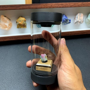 May include: A small, raw, multi-coloured mineral specimen featuring pink, green, and white hues, mounted on a square wooden base inside a clear cylindrical display container with black caps at the top and bottom, held in a hand.