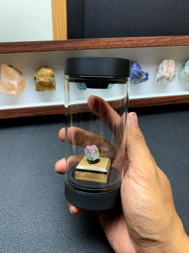 May include: A small, raw, multi-coloured mineral specimen featuring pink, green, and white hues, mounted on a square wooden base inside a clear cylindrical display container with black caps at the top and bottom, held in a hand.