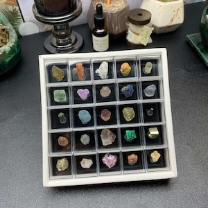 May include: A square white display case containing twenty-five individual clear plastic compartments, each holding a unique raw mineral specimen. The collection features various stones in shades of green, purple, yellow, clear, and metallic gold.