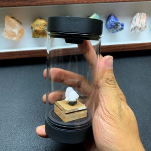 May include: A clear cylindrical display container with a black lid and base. Inside, a small white mineral specimen sits atop a square tan wooden pedestal. The container is held upright, showcasing the stone mounted on the central wooden platform.