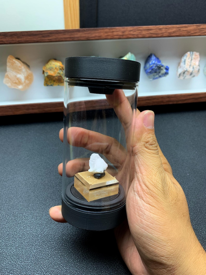 May include: A clear cylindrical display container with a black lid and base. Inside, a small white mineral specimen sits atop a square tan wooden pedestal. The container is held upright, showcasing the stone mounted on the central wooden platform.