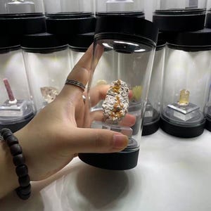 May include: A clear cylindrical display case with a black base and lid, featuring an integrated LED light at the top. Inside the container, a rough, textured mineral specimen with orange and white crystalline deposits sits on a small clear stand.