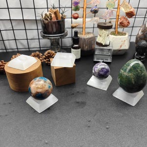 Puede incluir: Una colección de objetos decorativos que incluye varias esferas de piedra pulida y una piedra en forma de huevo, cada una sobre un soporte cuadrado esmerilado. También hay dos bloques de madera, dos árboles de alambre y una botella de vidrio.
