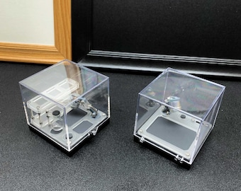 May include: Two square clear acrylic display cases with black bases. The left case contains a miniature white rectangular device with a small glass bottle. The right case holds a miniature grey rectangular device with a metallic mechanical arm.