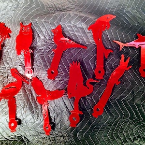 May include: A set of 13 red metal cutouts of various animals and objects, including an owl, a fish, a bird, a palm tree, a dolphin, a shark, a pelican, a crane, and a flag. The cutouts are all painted red and have a rustic, hand-crafted look.
