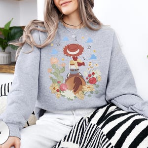 K&ouml;nnte beinhalten: Ein graues Sweatshirt mit Rundhalsausschnitt und einem Grafikdruck einer Cowgirl mit rotem Hut und geflochtenem Haar auf einem braunen Pferd. Das Motiv ist umgeben von bunten Blumen, gr&uuml;nen Kakteen, kleinen blauen Wolken und Noten im Hintergrund.