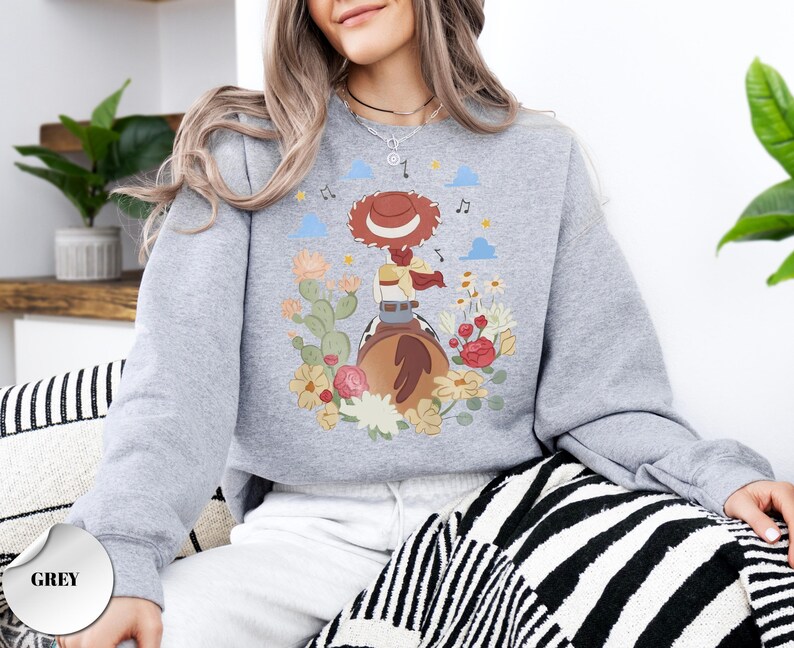 K&ouml;nnte beinhalten: Ein graues Sweatshirt mit Rundhalsausschnitt und einem Grafikdruck einer Cowgirl mit rotem Hut und geflochtenem Haar auf einem braunen Pferd. Das Motiv ist umgeben von bunten Blumen, gr&uuml;nen Kakteen, kleinen blauen Wolken und Noten im Hintergrund.